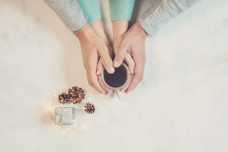 Couple in love holding hands with coffee on white marble table, with christmas lights. Photograph...