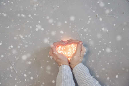 womans hand making shape of a heart, holding warm Christmas lights, wooden background, top view