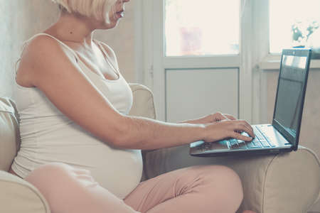 Young beautiful pregnant woman working on laptop. Pregnant business woman searching information and online shopping on laptop prepare for childbirth and parenting on sofa at homeの写真素材