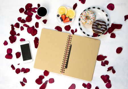 Morning Cup of coffee, empty notebook and roses flowers, fruits and donuts on white table from above. Cozy Breakfast. Flat lay style.の写真素材