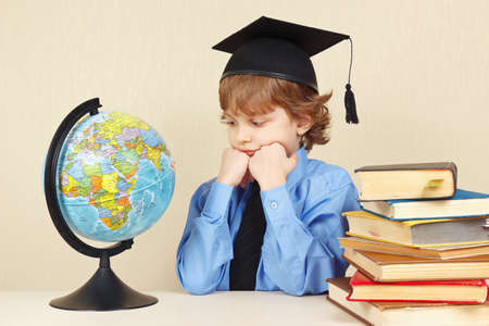 Little pensive boy in academic hat looks at a globe among the old booksの写真素材