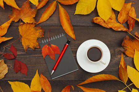 Cup of coffee, black notebook and pen among autumn fallen leaves on a wooden background. View from above.の写真素材