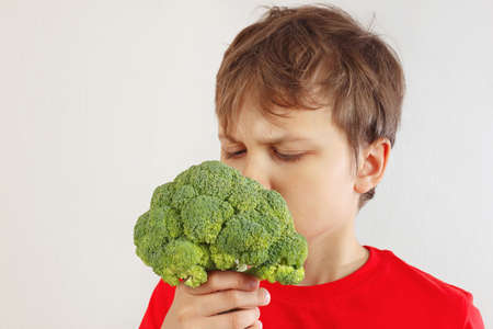 Little funny boy in a red shirt sniffing broccoli on a white backgroundの写真素材