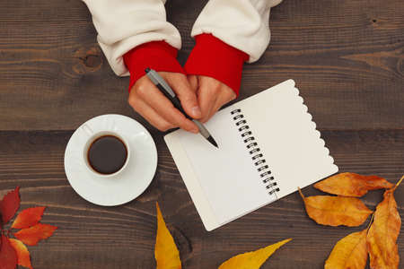 Hands of man with a pen and notebook at a wooden table with a cup of espresso and autumn leaves