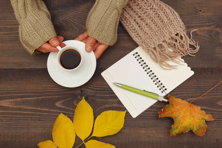 Hands of writer with a cup of espresso and scarf at wooden table with a notebook and pen and autumn leavesの写真素材
