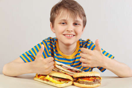Little funny boy in a striped shirt at the table recommends hamburgers and sandwich on a white backgroundの写真素材