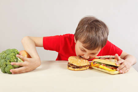 Boy in red at the table chooses between fastfood and vegetable and fruits on a white backgroundの写真素材