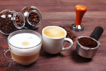 Cups of espresso and cappuccino, holder with ground coffee, tamper and cans of coffee beans on the table.の写真素材