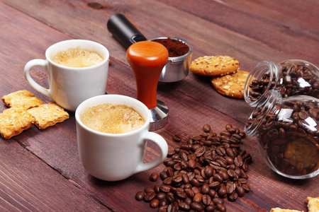 Two cups of espresso, crackers, holder with ground coffee, tamper and cans of coffee beans on the wooden table.の写真素材