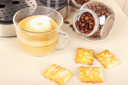 Cup of cappuccino under coffee maker, crackers and cans of coffee beans on the kitchen table.の写真素材