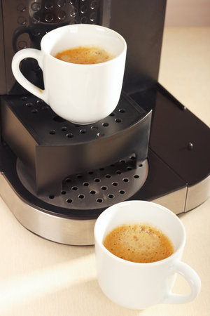 Two white cups of espresso under the coffee maker on the kitchen table.の写真素材