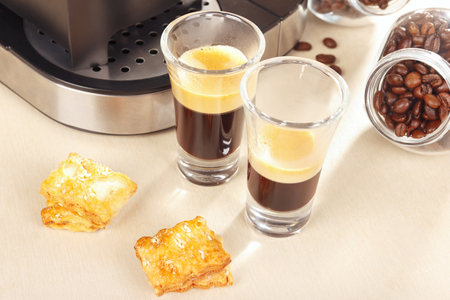 Two glass shorts of espresso near the coffee maker, crackers and cans of coffee beans on the light table.の写真素材