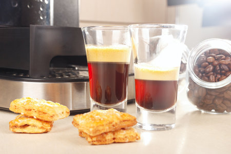 Two glass shorts of espresso near the coffee maker, cookies and cans of coffee beans on the kitchen table.の写真素材