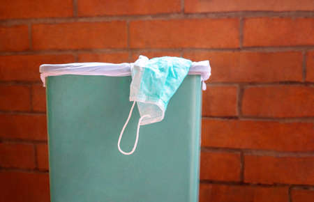 Medical mask used by sick african-american woman lies in green trash can against red brick wall....