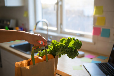 Close-up of woman holding paper bag with vegetables in kitchen at homeの素材