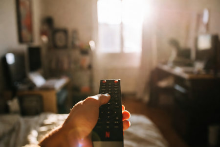 Hand holding TV remote control on blurred background of living room interior.の素材
