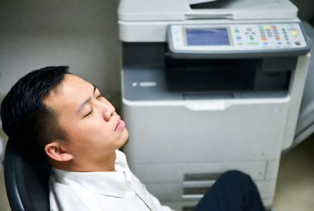 Young asian business man sleeping on office desk with copier in backgroundの素材