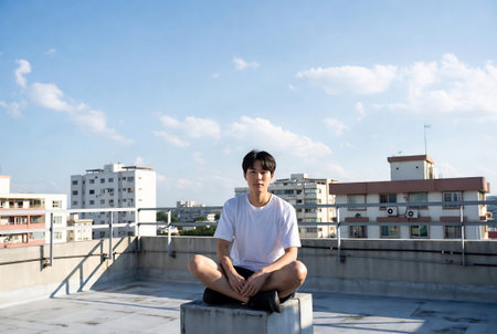 young asian man sitting on the roof of a building with blue skyの素材