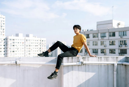 Handsome young man in yellow t-shirt and black pants sitting on the roofの素材