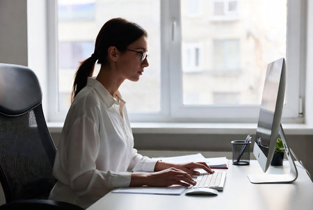 Side view of young businesswoman using computer while sitting at workplace in officeの素材