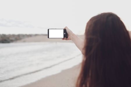 Young girl take selfie from hands with white blank smartphone on the beach, back view, selective focus.の写真素材