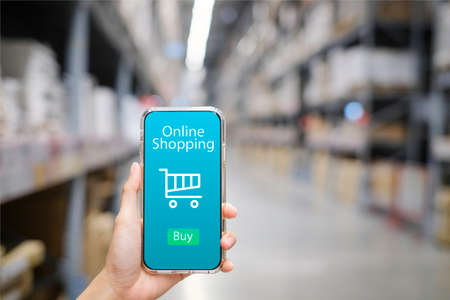 Smart phone online shopping in woman hand. Network connection on mobile screen. Payments online. Huge distribution warehouse with high shelves and loaders background.の写真素材