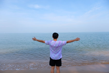 man on beach hold hands arms up, rear view guy wearing summer hat, standing back looking to sea blue sky horizon, vacation concept of freedom travel oceanの写真素材