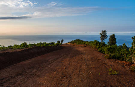 A mountainous dirt road overlooking the sea, a bumpy road with red soil amidst forest trees and blue sky facing the sea with sunset, wandering among the beautiful green nature.の写真素材