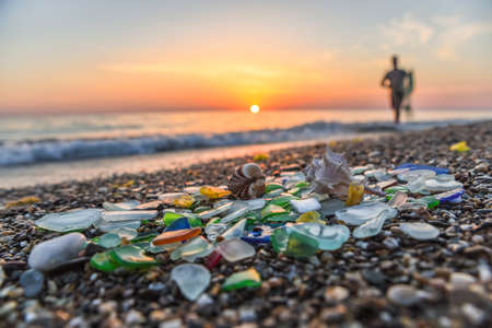 Collecting shells on beach at sunset, hobby of collecting seashells, sea glass, shells and pebbles in sandy beach and waves, in background and silhouette of young man collecting them, summer vacationの写真素材