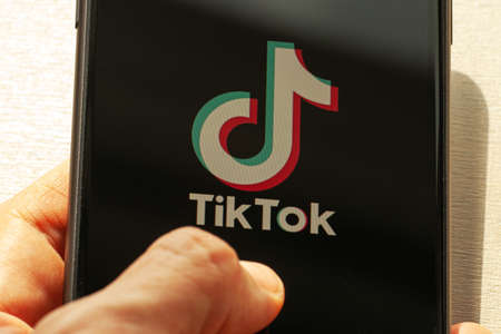 TikTok application icon on Apple iPhone 11 screen close-up. Hand holding smartphone Tik Tok icon on wooden background.  Tiktok Social media network from China.のeditorial素材