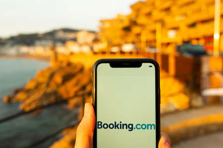 Hands holding iPhone 11 with an app of booking.com service. Holidays and vacation concept. A woman finding a place to stay or booking a hotel on a sunny day next to Mediterranean seaのeditorial素材