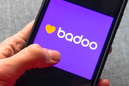 Young woman holding a smartphone with a Badoo dating app on screen. IPhone 11 with a social media chatting and meeting new people app logo.のeditorial素材