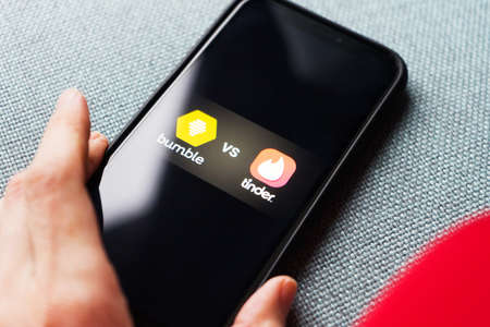 Young woman holding a smartphone with a Bumble vs Tinder dating icons on screen. IPhone 11 with a...