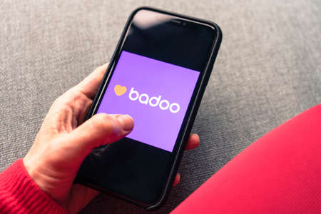 Young woman holding a smartphone with a Badoo dating app on screen. IPhone 11 with a social media chatting and meeting new people app logo.のeditorial素材