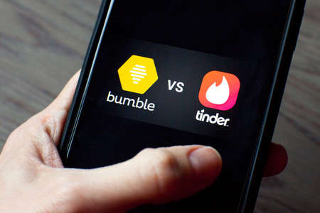 Young woman holding a smartphone with a Bumble vs Tinder dating icons on screen. IPhone 11 with a social media chatting and meeting people app logo.のeditorial素材