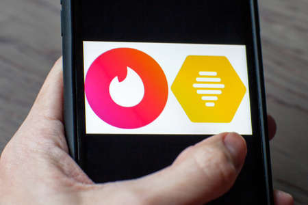 Young woman holding a smartphone with a Bumble vs Tinder dating icons on screen. IPhone 11 with a social media chatting and meeting people app logo.のeditorial素材