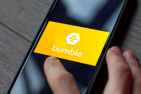 Hand holding a smartphone with a Bumble dating icon on screen. IPhone 11 with a social media app...