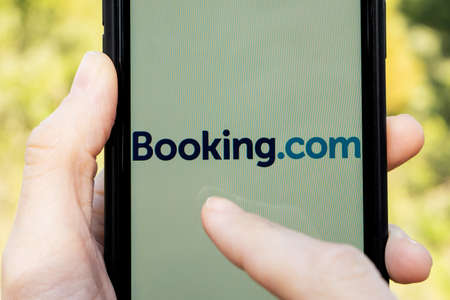 Hands holding iPhone 11 with an app of booking.com service. Holidays and vacation concept. A woman finding a place to stay or booking a hotel on a sunny day.のeditorial素材
