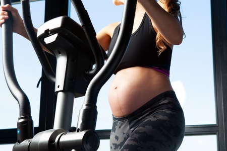 Pregnant woman in sports workout outfit training on an elliptical trainer . A close up of pregnant woman's belly. Healthy living and sports during pregnancy.の写真素材