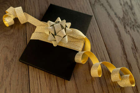 Close-up of present on a wooden vintage table. Black box with golden bow. Preparation for Christmas, birthday or other holidays and celebrations. Present packagingの写真素材