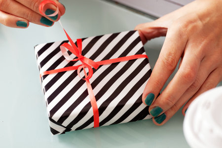 Female hands holding a gift wrapped in black and white stripes and tied with a red ribbon.