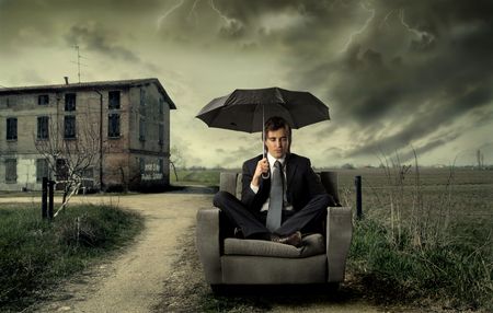 Businessman sitting under an umbrella on a sofa with countryside on the backgroundの写真素材