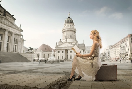Beautiful woman sitting on a suitcase and reading a book with monument in the backgroundの写真素材