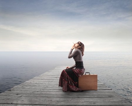 Beautiful woman sitting on a suitcase and smoking a cigarette with pier in the backgroundの写真素材