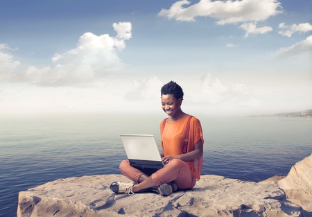Smiling african woman sitting on a rock over the sea and using a laptopの写真素材
