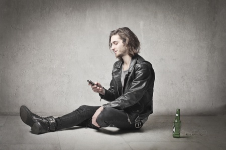 Young man in leather clothes uaing a mobile phone with a beer bottle beside himの写真素材