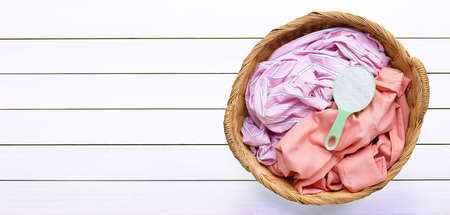 Colorful clothes with measuring spoon of detergent powder in laundry basket on white wooden...