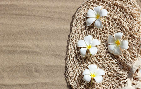 Summer background concept. Plumeria flowers on mesh beach bag on sand.