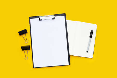 Blank a4 paper in black clipboard, black office paper clips with notebook and pen on yellow...