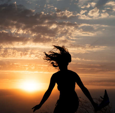 Young girl jumping silhouette with shawl on  beautiful cloudy sky with orange sunset and rays of lightの写真素材